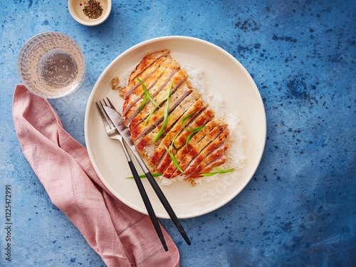 Breaded chicken is beautifully arranged on a bed of fluffy rice, topped with slices of green onion. A glass of water and a pink napkin complement the vibrant blue background photo