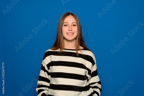 Horizontal portrait of a beautiful Hispanic teenage girl about 15s wearing a striped sweater smiling with braces on a blue background.