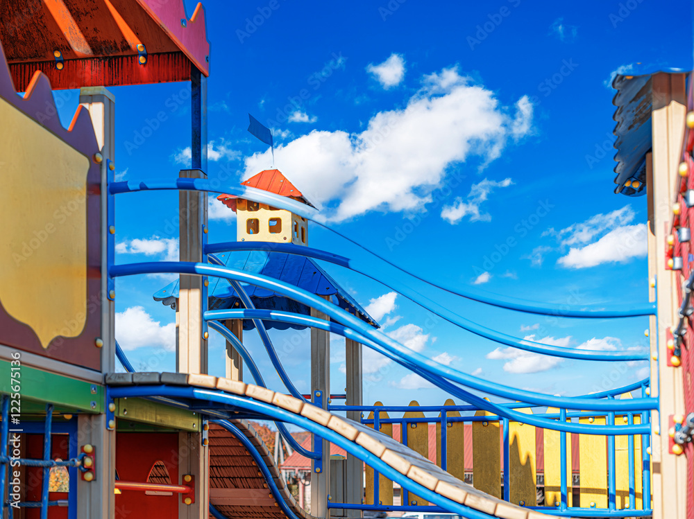 A colorful childrens playground structure with bridges, slides, and towers is set against a vibrant blue sky with fluffy clouds.