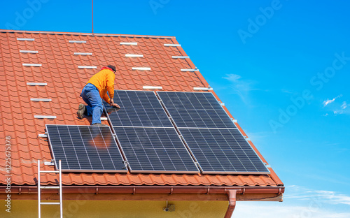Obraz na płótnie A worker is installing solar panels on the roof of a house covered with red tiles. The clear blue sky highlights the clean energy effort and rooftop solar panel technology. w salonie