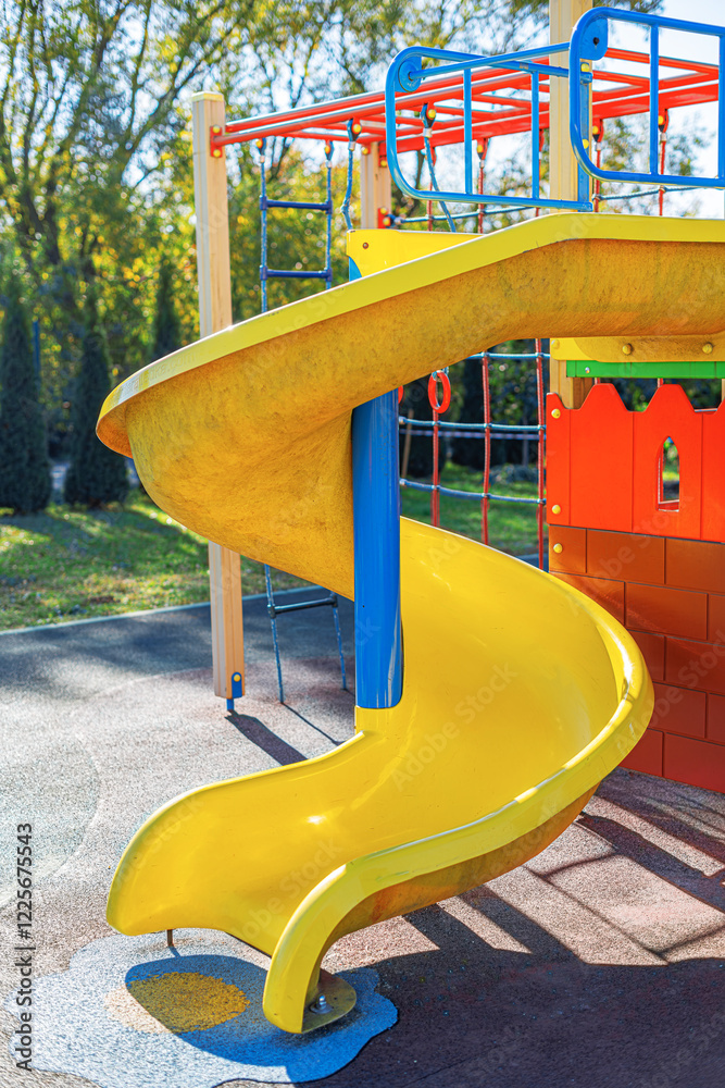 A vibrant yellow spiral slide stands in the playground, surrounded by bright blue metal railings and a colorful play structure.