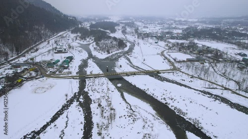 Aerial drone shot orbiting bridge over Lidder river at rafting point with small snow covered village homes in foreground and himalaya mountains and Srinagar Phelgam highway in distance photo