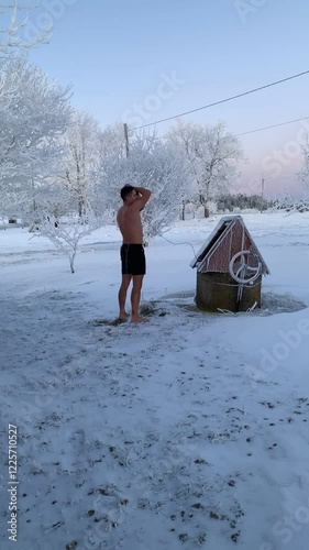 Vertical screen: A man in shorts pours a bucket of icy water over himself on a cold winter day. Steam rises from his body as the freezing water splashes, capturing a moment of resilience and refreshme photo