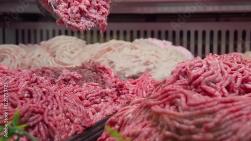 A panning close-up shot of a store's meat display featuring various types of fresh ground meat. An employee in black gloves uses a metal spatula to scoop minced meat from the display for packaging.