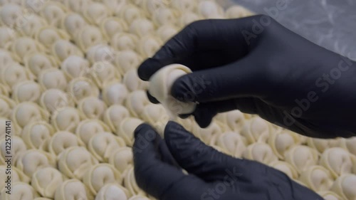 A person in black gloves wraps mincemeat in dough and molds a dumpling, then places it among the many molded dumplings lying in the background. Close-up shot.