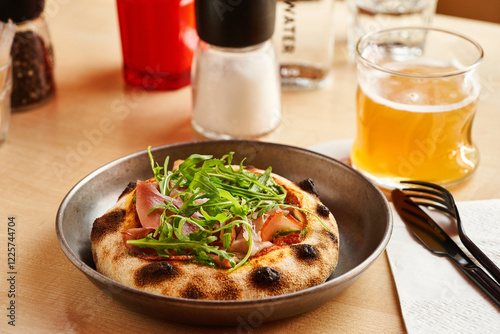 A freshly baked flatbread features savory prosciutto and vibrant arugula, complemented by a cold beer. The inviting atmosphere enhances the dining experience photo