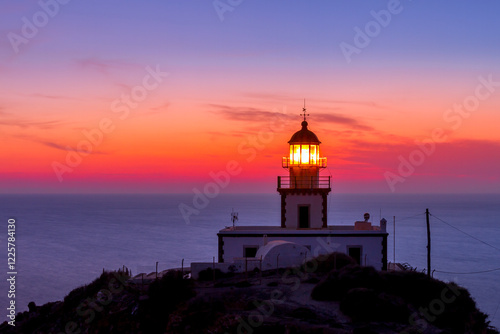 Fotografi The Akrotiri Lighthouse in Santorini, Greece, glows against a stunning sunset, casting its guiding light over the vast Aegean Sea—a breathtaking symbol of maritime history and island charm