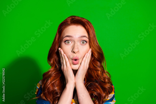 Young woman expressing surprise with hands on cheeks and colorful clothes against a vibrant green background