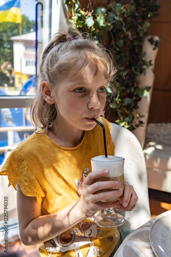 A little girl thoughtfully drinks cappuccino from a glass.
