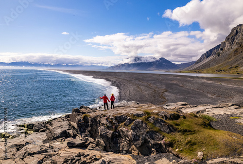 Scenic aerial drone view of couple in red jackets in Hvalnes Nature Reserve Black Beach and amazing mountains in background by the Atlantic Ocean in Iceland photo