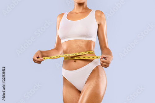 Perfect Body Concept. Cropped close up view of slim woman in white top and panties measuring her waist with yellow tape ruler. Unrecognizable lady checking waistline size isolated on blue studio wall