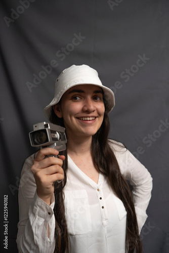 A cheerful woman with long hair smiles at the camera, wearing a white bucket hat mock up and white shirt, holding a vintage camcorder in a minimalist studio, radiating retro charm and creative energy. photo