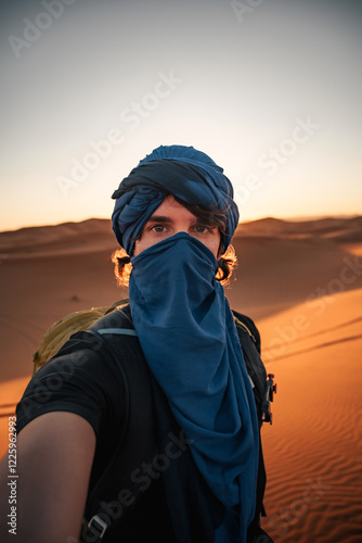 Backpacker taking a selfie while wearing blue turban and shemagh in the sahara desert at sunset photo