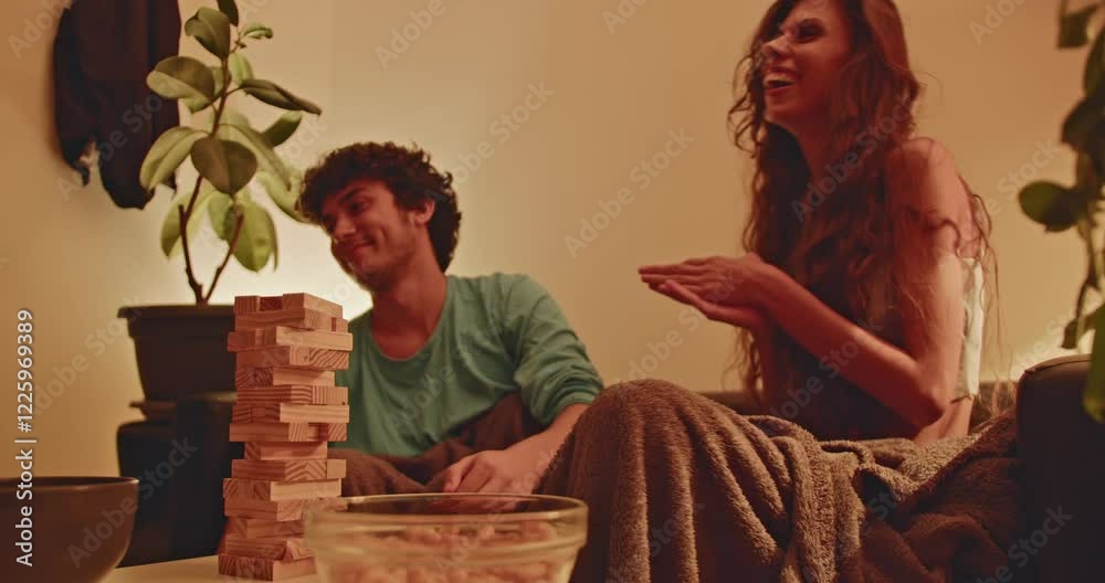 A young couple sits on a couch playing a game of Jenga. They laugh as they take turns pulling blocks from the tower.