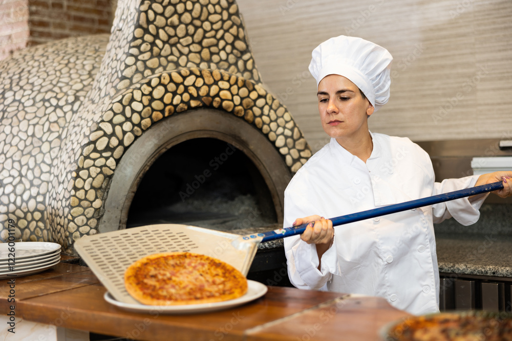 Portrait of smiling pizza chef at work, woman taking pizza from oven at restaurant kitchen