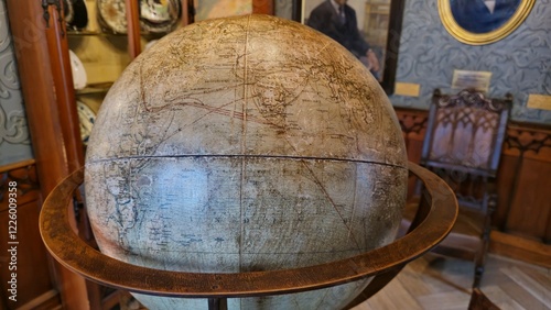 An antique terrestrial globe on a wooden stand, set in a historical room with ornate wooden furniture and portraits on the wall. The globe features detailed, weathered cartographic lines. photo