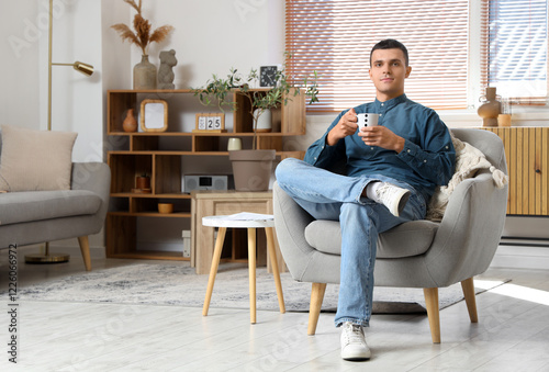 Handsome young man with cup of coffee sitting on armchair in modern living room