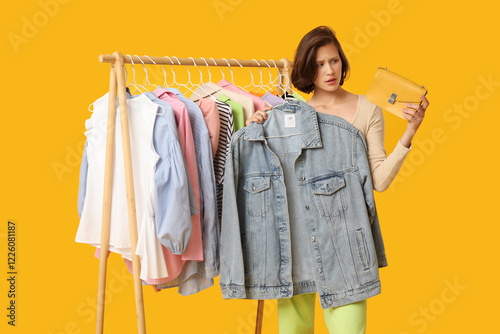 Thoughtful young woman near rack with different clothes choosing outfit on yellow background