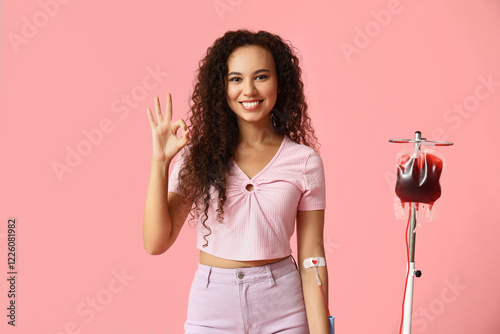 Young female African-American donor with applied medical patch donating blood and showing OK gesture on pink background photo