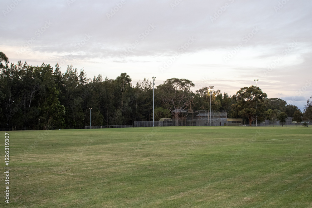 Obraz premium Captivating photo of a cricket ground in International Peace Park, Western Sydney, Australia. Stunning sunset glow through clouds, with lush greenery and trees in the background.