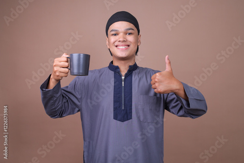 Portrait Of Young Asian Man Wearing Muslim Clothes Showing A Glass Of Water And Showing Thumb Up Isolated On Beige BackgroundPortrait Of Young Asian Man Wearing Muslim Clothes Showing A Glass Of Water photo