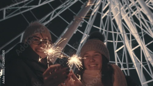 Happy Young Woman in Winter Clothes and Winter Hats Celebrate Christmas with Sparklers, standing together on the street with the background of a luminous Ferris Wheel a Christmas tree 4K Footage