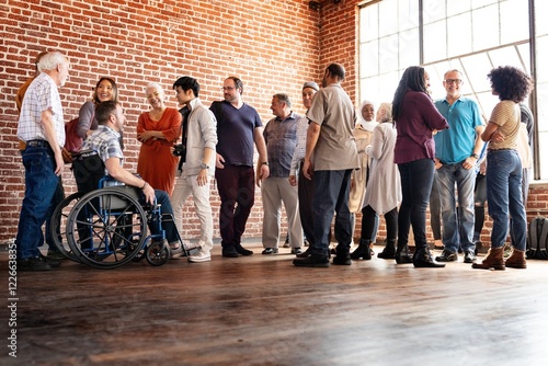 Diverse group of people socializing indoors. Men and women of various ethnicities and ages, including a person in a wheelchair, gathered in a brick-walled room.
