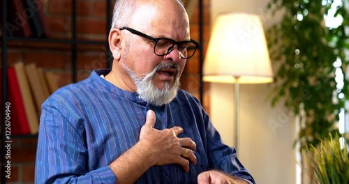 Indian senior retired man feels breathless from an asthma attack, uses inhaler to regain breath while sitting at his home desk