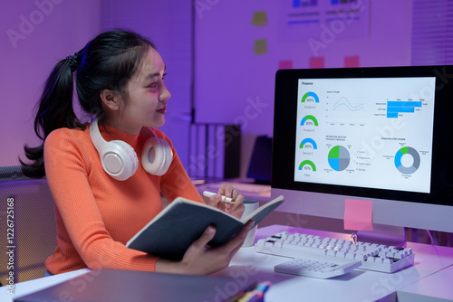 Focused young woman with headphones analyzing business data on a computer screen. She takes notes in a notebook, surrounded by a modern office setup with colorful charts