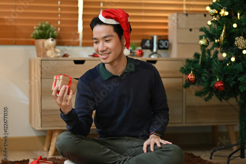 A young man in a Santa hat sits by a Christmas tree, smiling as he holds a small wrapped gift. The cozy holiday setting includes warm lights and festive decorations photo