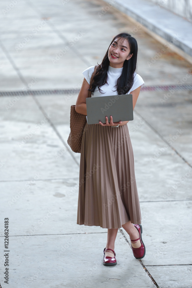 Fototapeta premium Asian businesswoman standing outdoors in elegant casual wear with pleated skirt and comfortable shoes. Working on laptop while carrying tote bag. Modern lifestyle concept of remote work and mobility