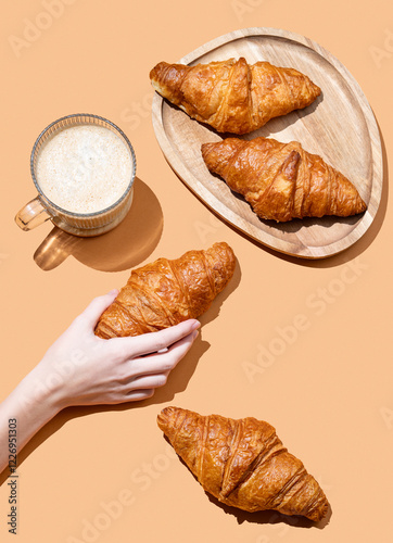 Hand holding fresh croissant on orange background with cappuccino cup and fresh pastry