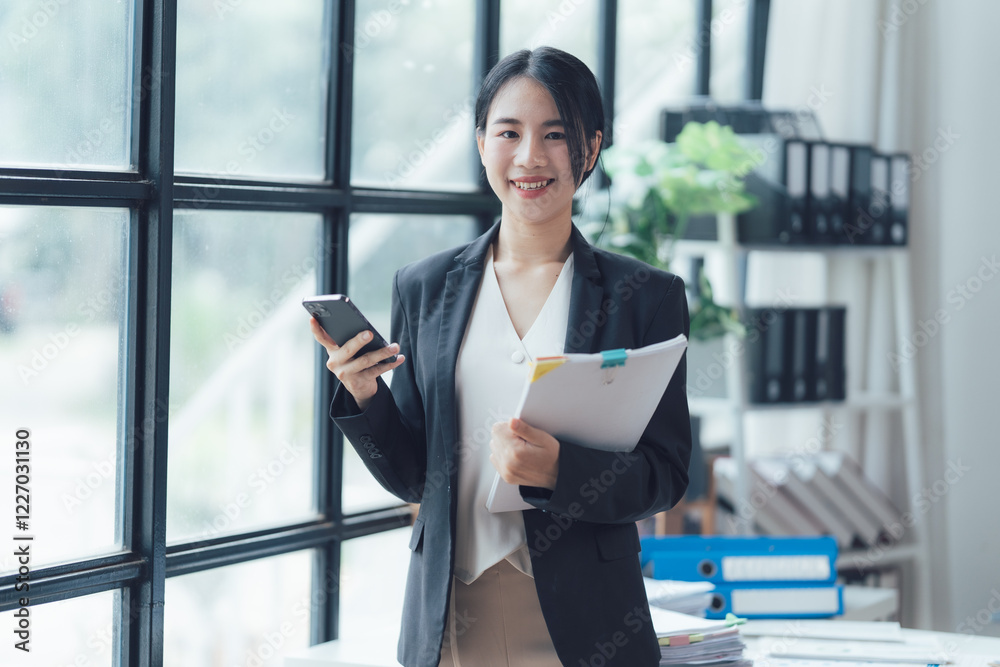 Businesswoman Confidence: A confident, smiling businesswoman in a sleek blazer holds a smartphone and documents against a backdrop of large windows, exuding professionalism and success. Businesswoman Confidence: A confident, smiling businesswoman in a sleek blazer holds a smartphone and documents against a backdrop of large windows, exuding professionalism and success.