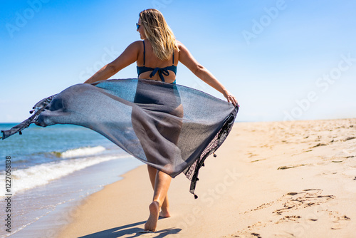 Beautiful middle aged woman holding scarf fluttering in wind walking on seashore on sandy beach. Back view	 photo