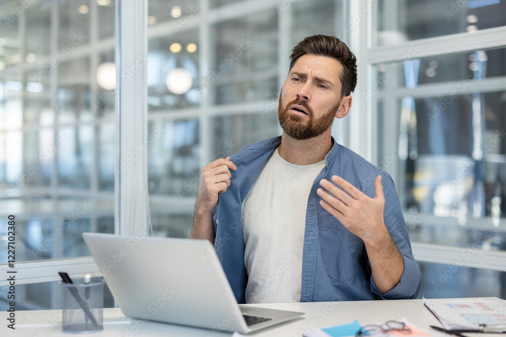 Heat in the office, broken air conditioner. Man trying to cool down at workplace with laptop, waving hand in frustration, lack of fresh air. Heat in the office, broken air conditioner. Man trying to cool down at workplace with laptop, waving hand in frustration, lack of fresh air.