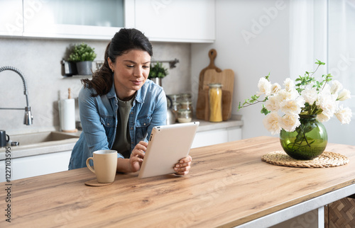Smiling young successful woman drinking coffee and using tablet computer while sitting in kitchen. Businesswoman reading emails at morning in her home. Remote work business owner female