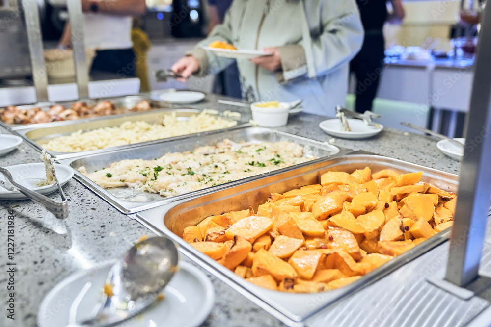 Fototapeta premium Close-up of a metal container with baked hearty food. A woman puts food in a cup on a buffet table in a restaurant. With space to copy. High quality photo