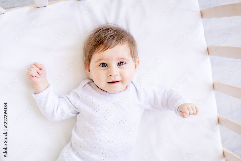 a small child close-up on a white cotton bed at home is lying on a split in a white bodysuit and smiling, a baby girl with big eyes of six months, top view a small child close-up on a white cotton bed at home is lying on a split in a white bodysuit and smiling, a baby girl with big eyes of six months, top view