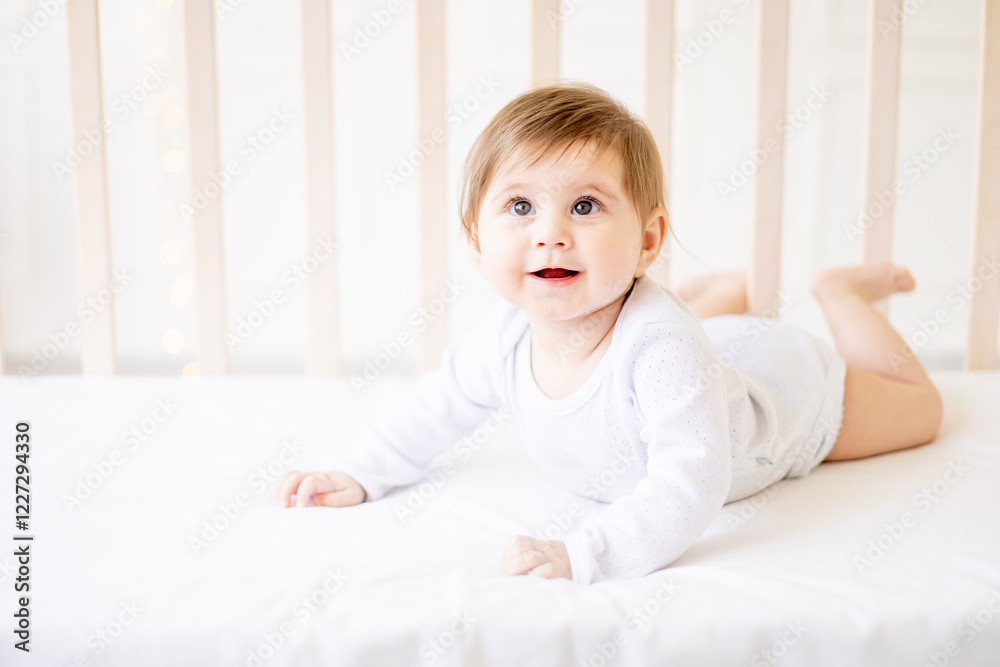 a small child close-up on a white cotton bed at home lies on her tummy and smiles, a baby girl with big eyes of six months a small child close-up on a white cotton bed at home lies on her tummy and smiles, a baby girl with big eyes of six months
