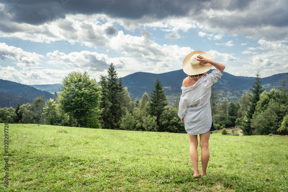 Young woman in hat standing on green meadow in mountains, rear view Young woman in hat standing on green meadow in mountains, rear view