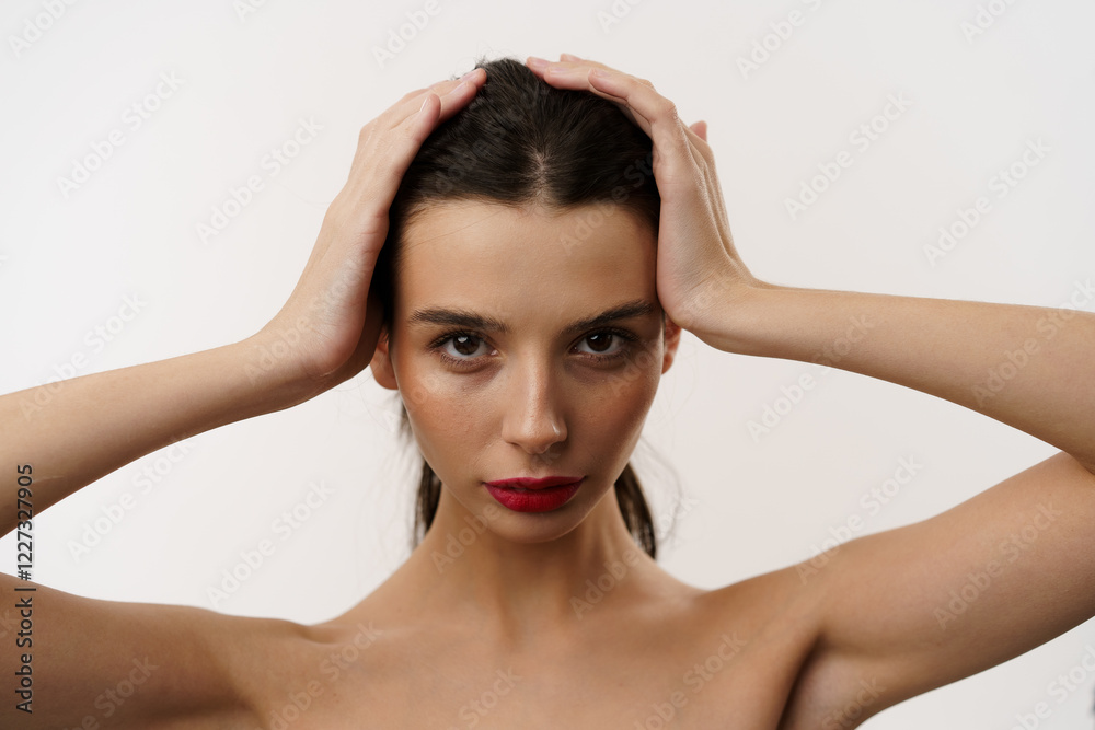 Fototapeta premium Young brunette woman with red lipstick posing against a white background, expressing emotions, and touching her face with her hands. Minimalistic beauty and fashion concept
