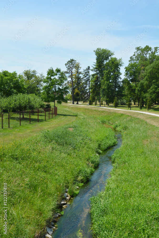 A stream flows through a lush green meadow in Erdody Park, Jastrebarsko, Croatia, under a bright blue sky with a path and trees lining the edges of the park A stream flows through a lush green meadow in Erdody Park, Jastrebarsko, Croatia, under a bright blue sky with a path and trees lining the edges of the park