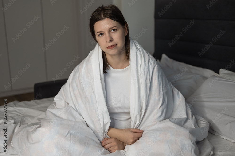 Young woman sitting on bed wrapped in white blanket looking thoughtful introspective. Broken sad woman looking straight into camera. personal problems. mental crisis. being trapped in your thoughts Young woman sitting on bed wrapped in white blanket looking thoughtful introspective. Broken sad woman looking straight into camera. personal problems. mental crisis. being trapped in your thoughts