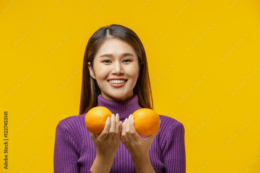 Beautiful young Asian woman with long brown hair wearing purple sweater smiling and holding two oranges in hands isolated on yellow background