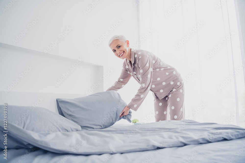 Elderly woman in polka dot pajamas making bed in sunlit bedroom Elderly woman in polka dot pajamas making bed in sunlit bedroom