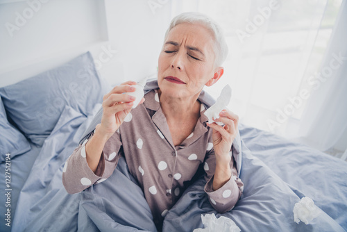 Elderly woman in polka dot pajamas feeling unwell sitting on bed holding tissue in morning light photo