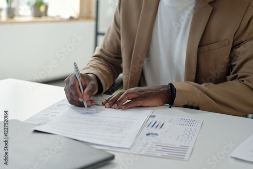 Hands of young unrecognizable male economist with pen reading business contract and signing it by desk