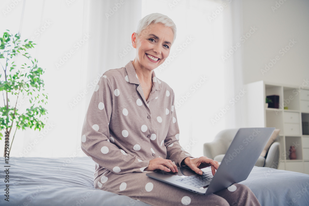 Elderly woman in pajamas smiling with laptop on bed in cozy bedroom Elderly woman in pajamas smiling with laptop on bed in cozy bedroom