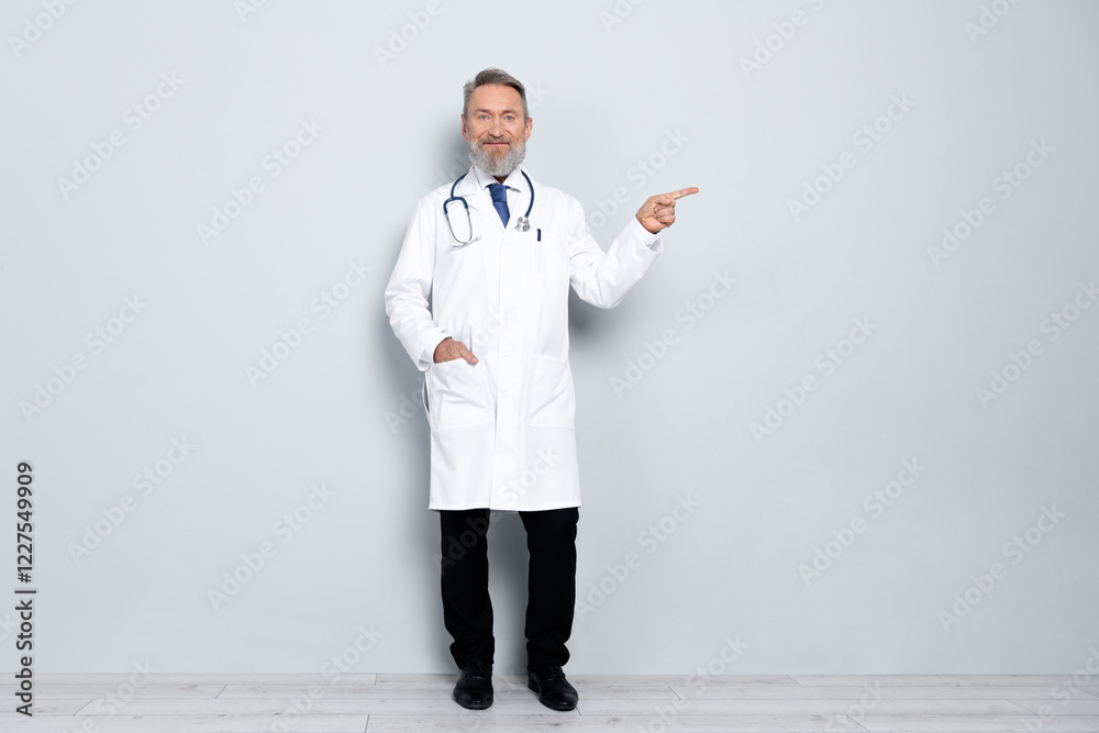 Senior male doctor wearing a white lab coat pointing outward with a friendly gesture on a light grey background Senior male doctor wearing a white lab coat pointing outward with a friendly gesture on a light grey background