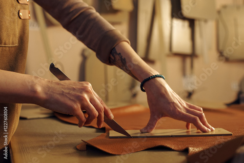 Artisan carefully cutting leather with sharp tool, focusing on detailed work. Engaging in precise manual process within well-organized workspace photo